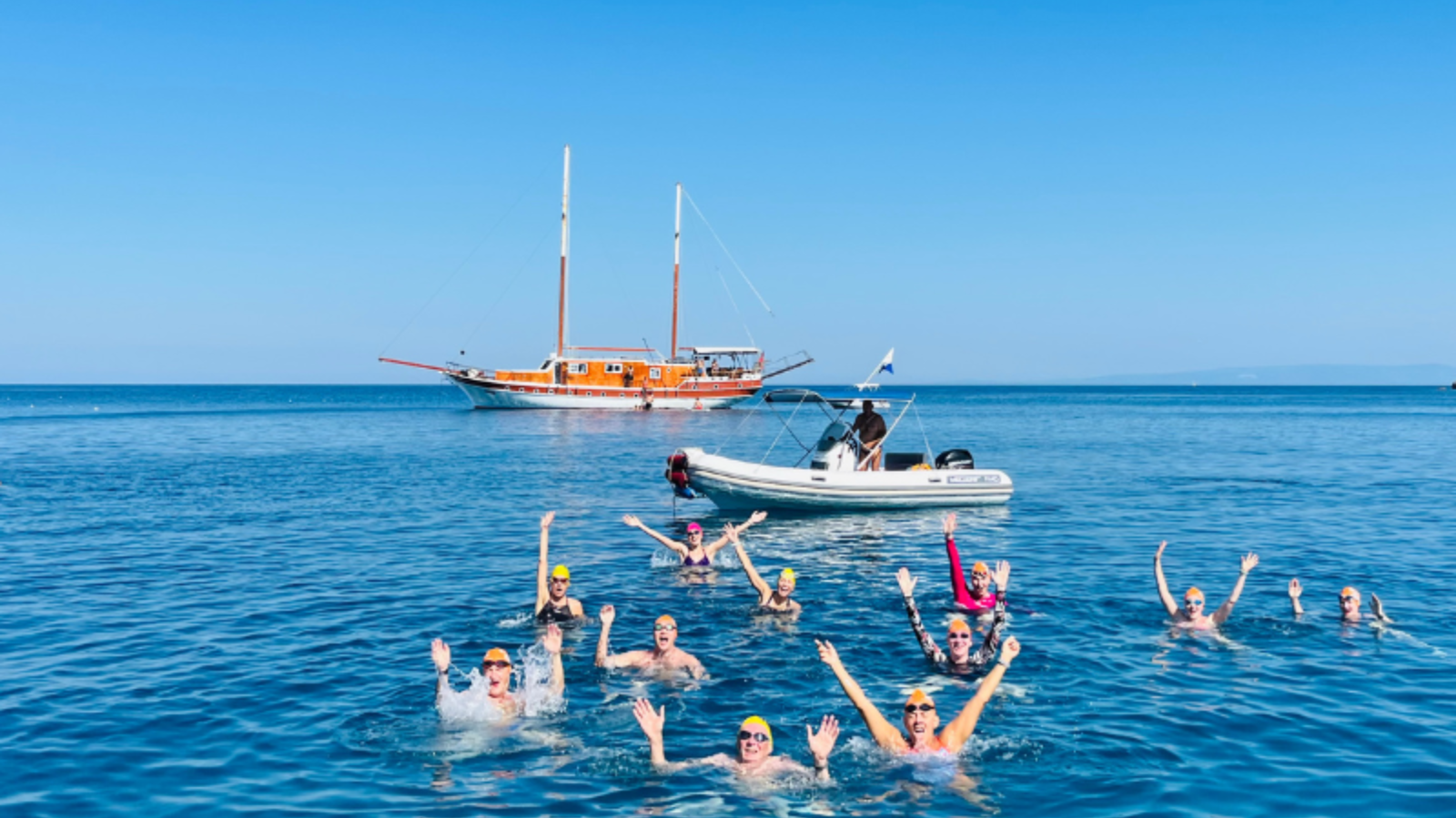 Group of open-water swimmers waving with both arms in a bright blue sea beside a sailing boat