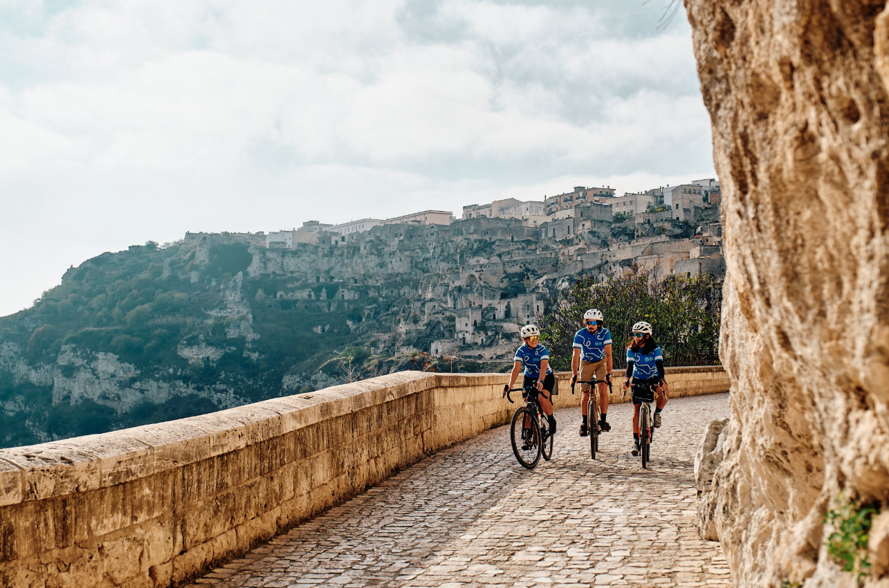 Three cyclists riding a cobbled road below an ancient hillside town in the afternoon light